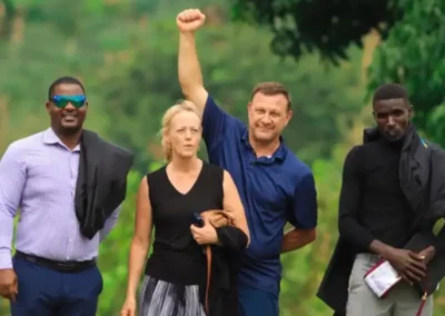 A group of four individuals stands outdoors, one with a raised fist, against a backdrop of green foliage