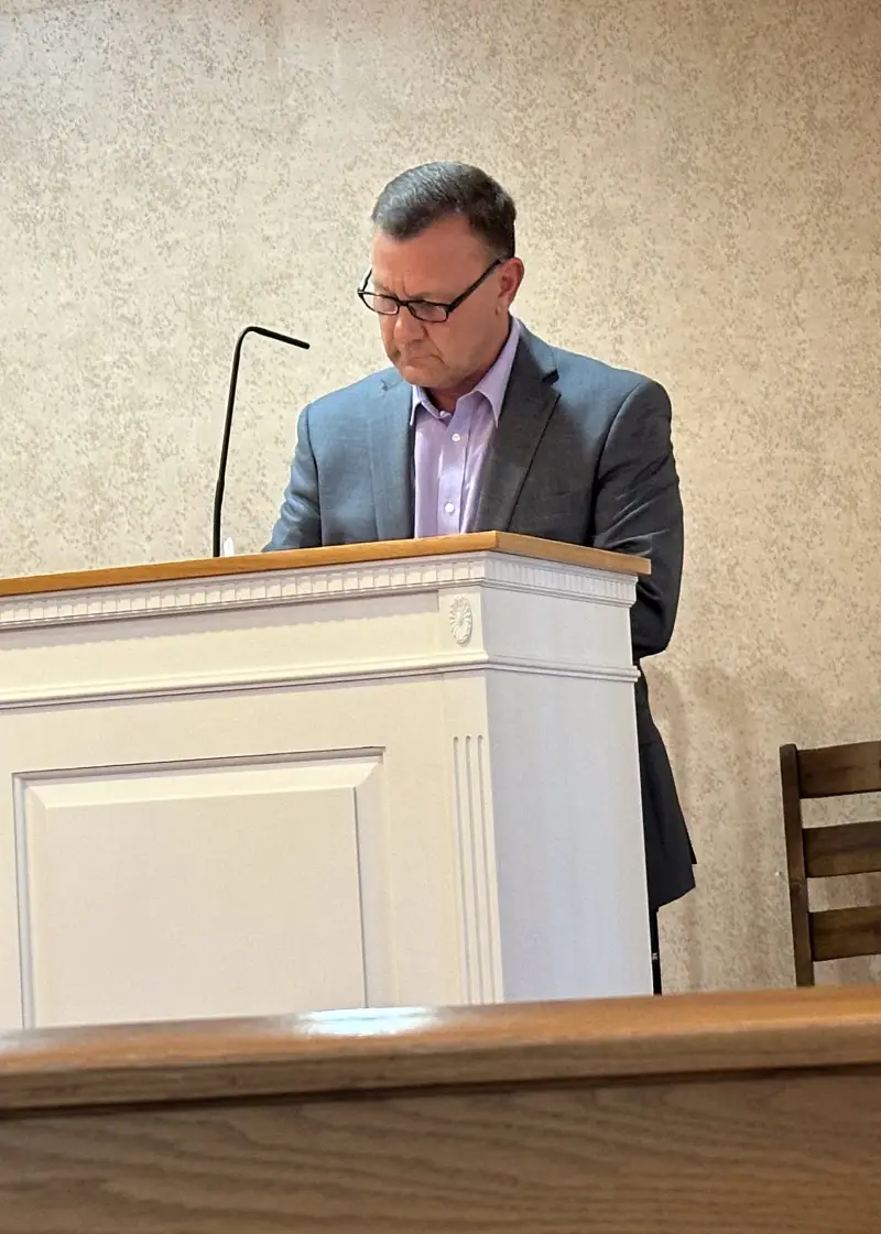 A person in formal attire stands at a podium in a modest room, delivering a speech, with a wooden chair in the background